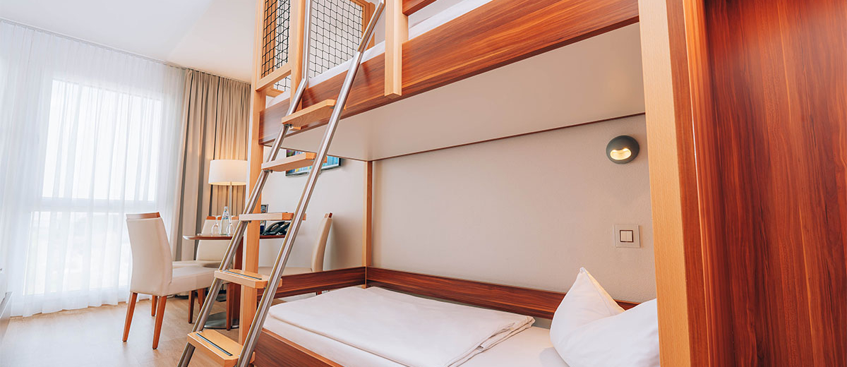 Hotel Victory Therme Erding Bellezza Seitliche Ansicht von einem Stockbett und einer Leiter ins obere Bett. Im Hintergrund ein Tisch mit Stühlen